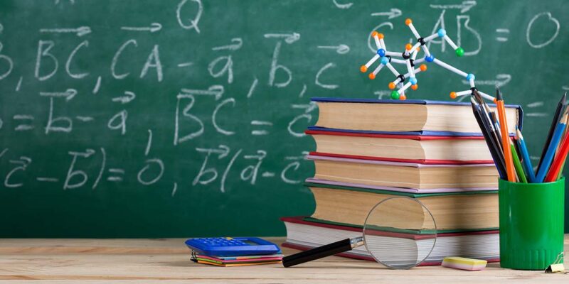 teachers desk with stack of books and chalkboard