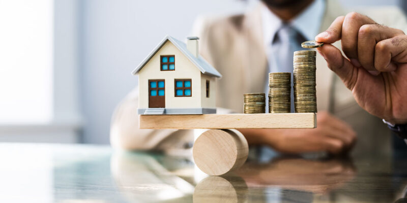 Man stacking coins on one side of the scale while the other side has a toy house